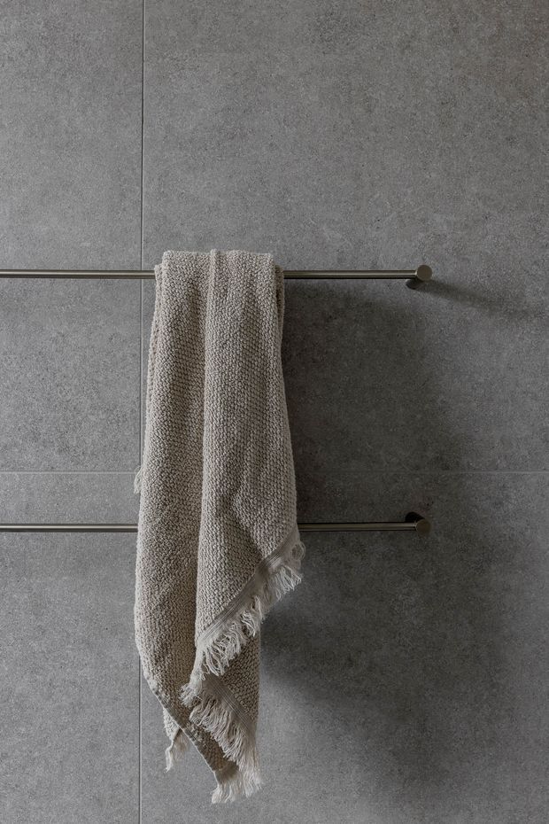 Close-up of a textured neutral towel hanging from two slim horizontal metal towel rails mounted on large grey stone-look wall tiles. The image has a restrained, monochromatic bathroom aesthetic with soft natural texture and minimal detailing.