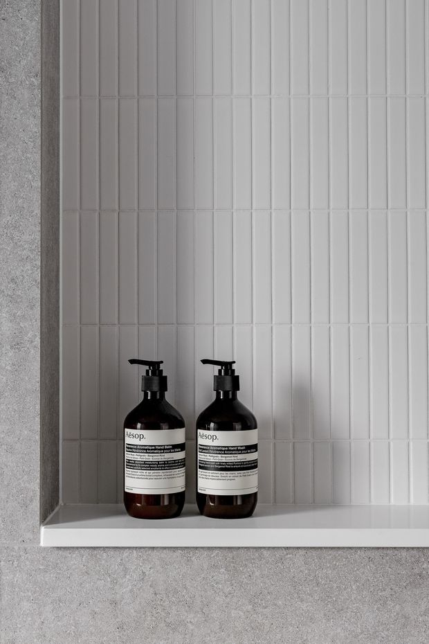 Close-up of a minimalist tiled wall niche with a white ledge, holding two dark amber pump bottles. The niche is framed by grey textured surfaces, and the wall is finished with narrow vertically oriented white tiles in a clean, repetitive grid.