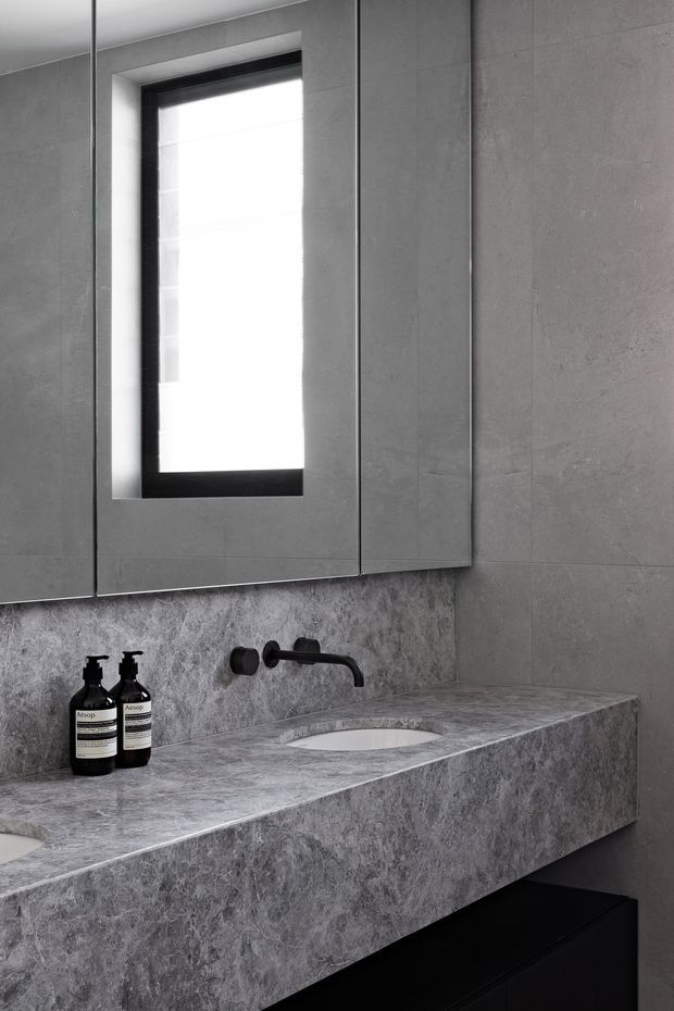 A close-up view of a refined contemporary bathroom vanity. The space features a large grey stone-look benchtop and integrated basin, dark matte wall-mounted tapware, grey tiled walls, a black-framed window, mirrored cabinetry above the vanity, and dark under-vanity joinery. The monochrome palette and clean lines create a minimalist, moody atmosphere.