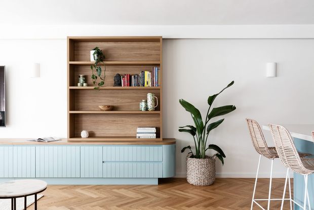 Bright contemporary living and dining area featuring a built-in timber shelving unit with decorative objects and books above a pale blue fluted media cabinet. The room has white walls, herringbone timber flooring, woven bar stools at a pale blue kitchen or dining counter, and a large indoor plant in a textured basket planter.