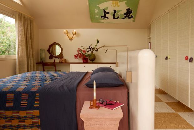 A warm, eclectic bedroom with a vintage-inspired dressing table and oval mirror, patterned blue and orange bedding, a pink bedspread and bedside table, brass wall sconce, floral arrangement, and a large Japanese-style wall hanging. The room has cream walls, built-in wardrobes, tiled flooring, and soft natural light from a curtained window.