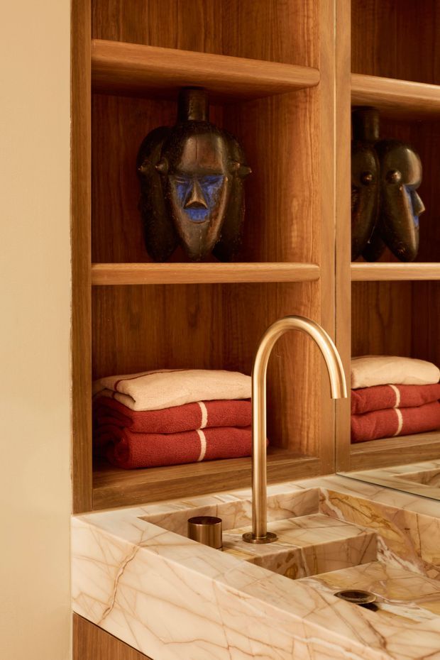 Close-up of a richly detailed bathroom vanity with a polished veined stone countertop and integrated basin, featuring a warm wood open shelving niche, folded red and cream towels, sculptural dark decorative objects, and a large mirrored panel. A curved brass-gold tap and matching drain and cup accents add a refined, warm luxury feel.
