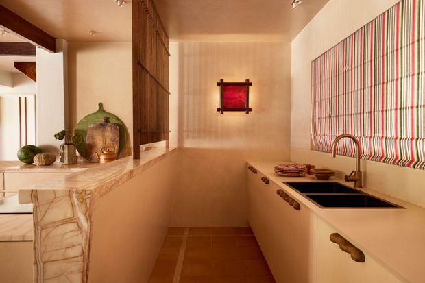 Warmly lit galley kitchen with cream cabinetry, dark timber accents, a pale marble or stone peninsula, terracotta-toned tiled flooring, and a double black undermount sink beneath a bold vertically striped Roman blind. A pass-through or adjoining area is visible at left, with decorative objects on the benchtop and a small red wall artwork.
