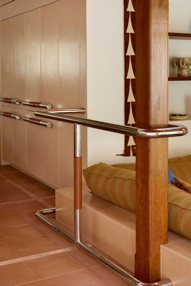 Close-up interior view of a mid-century-inspired living area with a polished tubular metal safety railing featuring leather-wrapped handrail sections. The railing borders a stepped or sunken seating area with warm striped upholstery. A vertical timber post, built-in shelving, patterned wall textile, and large terracotta-toned floor tiles create a warm retro character.