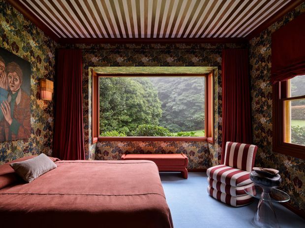 A richly decorated bedroom with dark floral patterned wallpaper, deep red curtains, a large timber-framed picture window overlooking lush greenery, and a dramatic striped coffered ceiling. The room includes a large dusty-rose upholstered bed, matching ottoman, bold red-and-white striped lounge chair, and sculptural side table. The overall look is theatrical, eclectic, and highly saturated, with a strong vintage character.