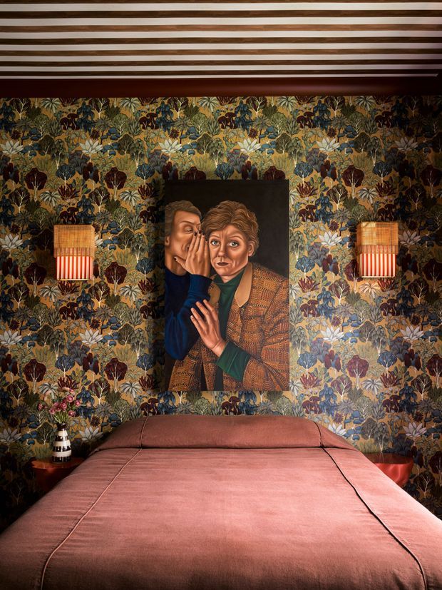 A richly styled bedroom with a bed dressed in dusty rose-pink bedding, surrounded by dark botanical patterned wallpaper. A large figurative artwork hangs above the bed, flanked by matching wall sconces with woven shades and striped lower panels. The ceiling features exposed or decorative timber slats, creating a dramatic, intimate and highly layered interior.