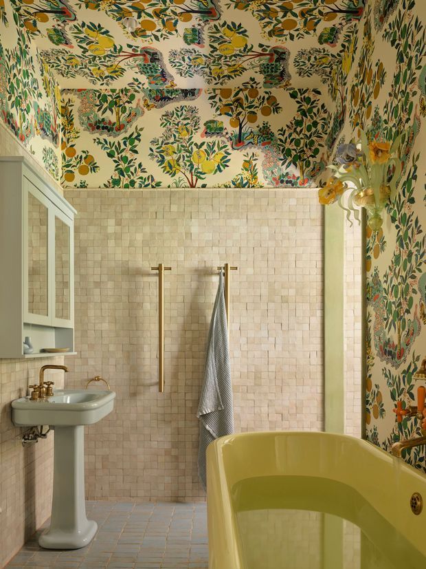 Eclectic, highly decorative bathroom featuring a freestanding pale yellow-green bathtub, pedestal basin with brass tapware, a wall-mounted mirrored cabinet, and a brass-toned heated towel rail. The lower walls are clad in small pale stone tiles, while the upper walls and ceiling are covered in a vivid botanical and bird-patterned wallpaper. The space has warm natural colours, ornate detailing, and a playful maximalist character.