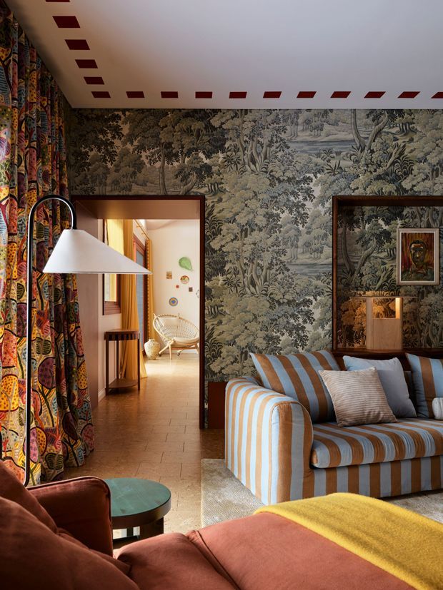Eclectic living room with richly patterned botanical wallpaper, bold multicoloured curtains, and a blue-and-white striped sofa. A large sculptural floor lamp, framed artwork, timber-framed openings, warm tiled flooring, and layered upholstered seating create a colourful, refined and theatrical interior. A secondary seating area is visible through the doorway.