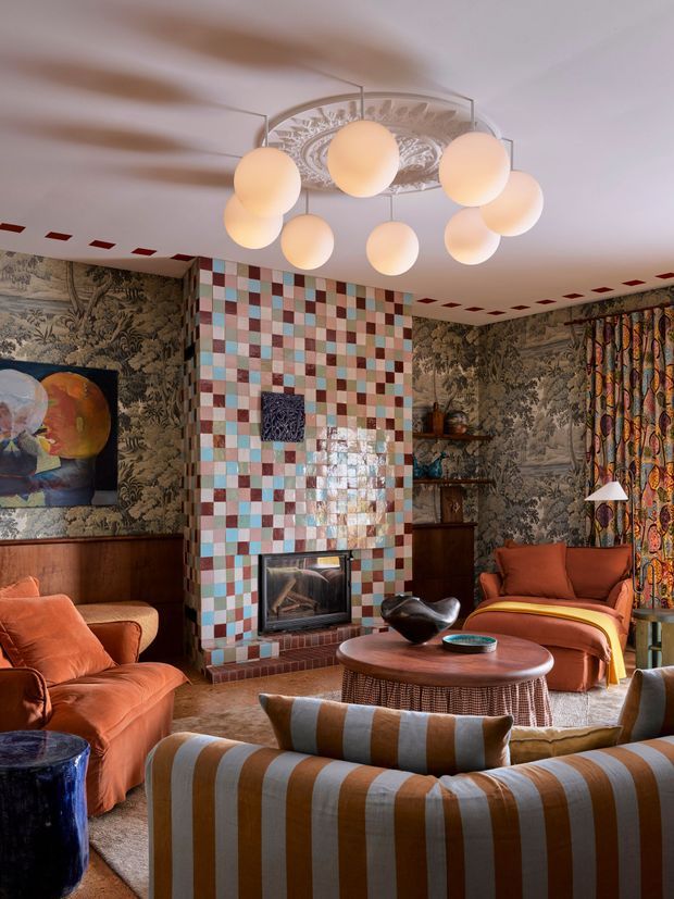 Eclectic, richly layered living room featuring patterned botanical wallcoverings, a colourful checkerboard tiled fireplace surround, warm terracotta and ochre upholstered seating, a rounded woven coffee table, and a sculptural multi-globe pendant light. Decorative shelving, artwork, patterned curtains, and bold textiles create a theatrical, maximalist atmosphere.