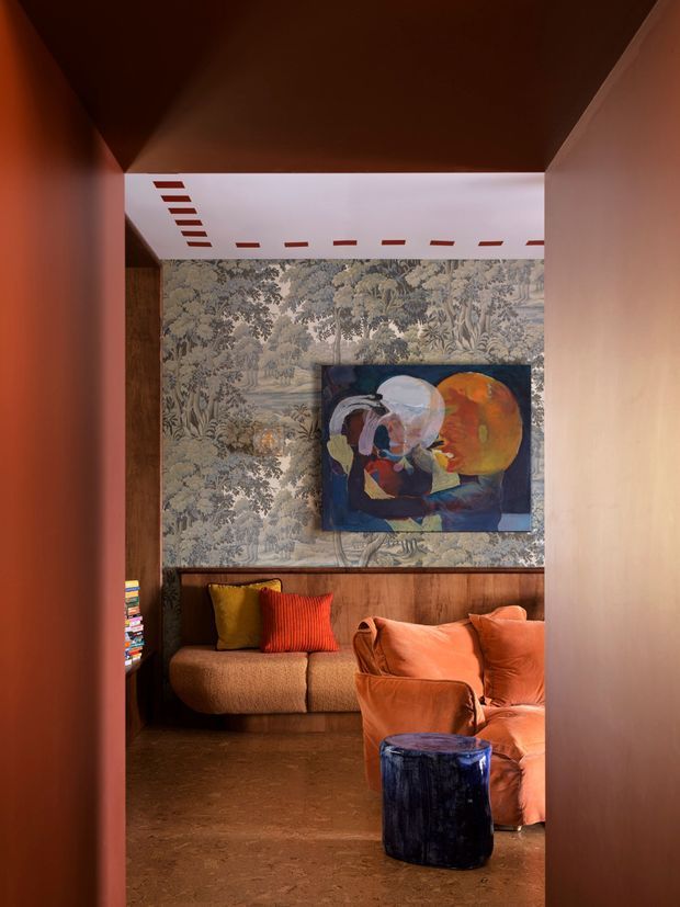 A richly layered, intimate sitting area viewed through a narrow passage, with warm terracotta-painted walls and ceiling, a patterned botanical wallpaper feature wall, dark timber wainscoting, and a brown floor. A tan sofa with mustard and red cushions sits beneath a colourful abstract artwork, while a burnt-orange armchair and ottoman occupy the foreground. The space has dramatic warm lighting and a curated, eclectic character.