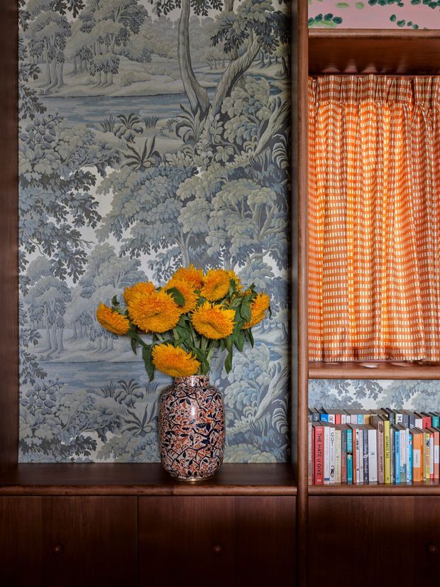 Close-up interior vignette featuring dark timber built-in cabinetry and shelving, with a blue-grey pastoral botanical wallcovering behind a decorative vase of orange flowers. The shelving includes a bright orange patterned fabric panel and a small collection of books, creating a richly layered, traditional and eclectic decorative composition.