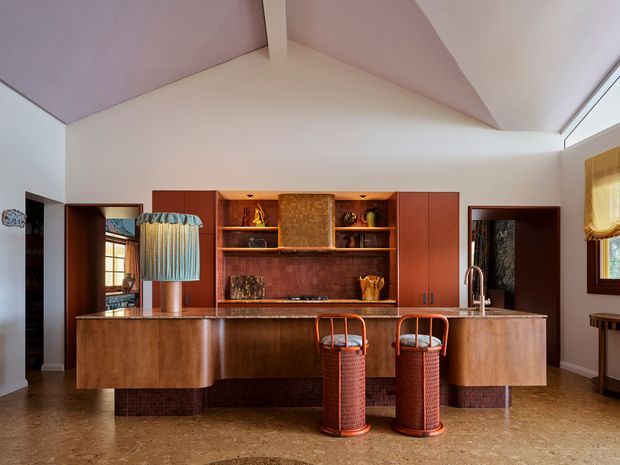 A warm, richly detailed kitchen with a large curved timber island and integrated seating, dark reddish timber cabinetry, open shelving, and a central textured backsplash. Two woven bar stools sit at the island, while patterned pendant lighting, decorative objects, and windows add character. The high angled ceiling and clerestory-style glazing bring in natural light, and the overall palette is earthy and refined.