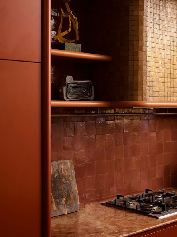 Close-up view of a richly coloured kitchen with deep terracotta and reddish-brown tiled walls, integrated open shelving, rounded terracotta-toned cabinetry and rails, and a gas cooktop set into a mottled stone benchtop. Warm, low lighting and dark shelving create a dramatic, intimate atmosphere. The image focuses on fixed kitchen joinery, backsplash tiling and cooking fixtures, with a few small kitchen accessories on the shelves and benchtop.