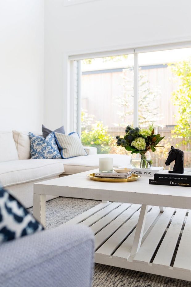 Bright, airy living room with a white sectional sofa, patterned blue-and-white cushions, and a large white slatted coffee table. A floral arrangement, candle, books, and decorative horse sculpture sit on the table. Large windows overlook a leafy fenced yard, creating a fresh, relaxed coastal-inspired atmosphere.