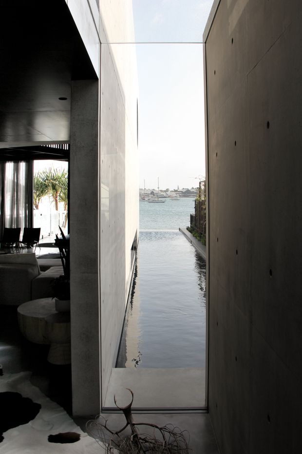 A dramatic narrow architectural view through a full-height glazed opening between tall raw concrete walls, framing a long reflecting pool that leads toward a calm waterfront and harbour beyond. A dim contemporary living area with dark finishes, seating, a round low table, and indoor planting is visible to the left. The composition emphasises strong vertical planes, water, and natural light.