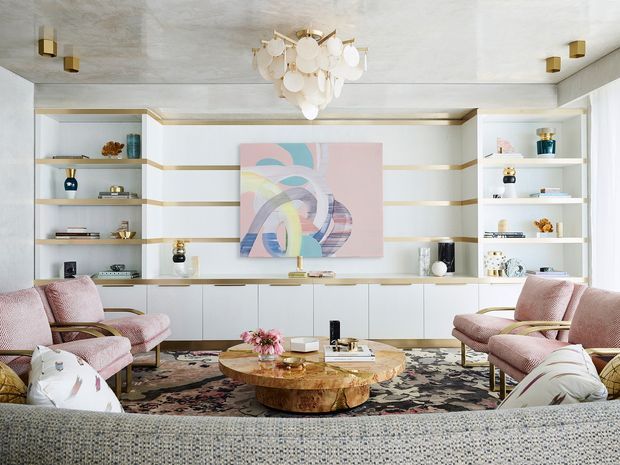 A refined living room with a built-in white wall unit combining open display shelving and low cabinetry, accented by thin brass-gold trim. The room features blush-pink upholstered lounge chairs, a large textured sofa in the foreground, a sculptural round wood coffee table, patterned area rug, abstract pastel artwork, and a statement pendant light with layered white discs. The overall scheme is bright, elegant, and softly contemporary with glamorous traditional influences.