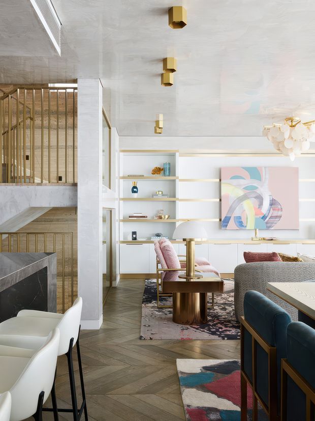 Bright, refined contemporary open-plan living and dining area with a white plastered ceiling, pale stone-clad structural column, built-in white shelving and cabinetry with brass trim, and herringbone timber flooring. The room includes a dining table with white upholstered chairs, a sculptural chandelier, a coffee table, upholstered lounge seating, colourful abstract artwork, and patterned rugs. A timber and brass balustrade at left suggests a stair or split-level connection. The styling combines polished metallic accents, soft pink and blue upholstery, and curated decorative objects.