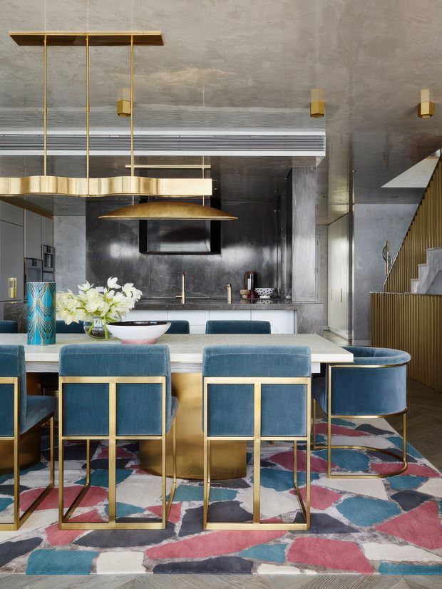 A dramatic open-plan dining and kitchen interior with a long dining table surrounded by deep blue upholstered chairs in brass-gold frames. A sculptural linear pendant and large dome pendant hang above the table. The kitchen features dark reflective cabinetry or wall panels, a pale island, integrated appliances, and a metallic rangehood. Concrete-look ceiling and walls, a patterned multicolour rug, brass detailing, and a staircase with vertical timber slats create a luxurious, highly layered interior.