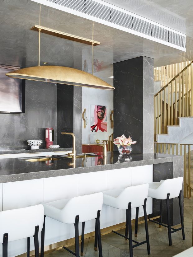 A sophisticated kitchen and bar area with a dark marble or stone-clad backsplash and island, white cabinetry, brass accents, and a large sculptural pendant light. White upholstered bar stools line the island, while a gold-toned stair balustrade and staircase are visible behind the kitchen.