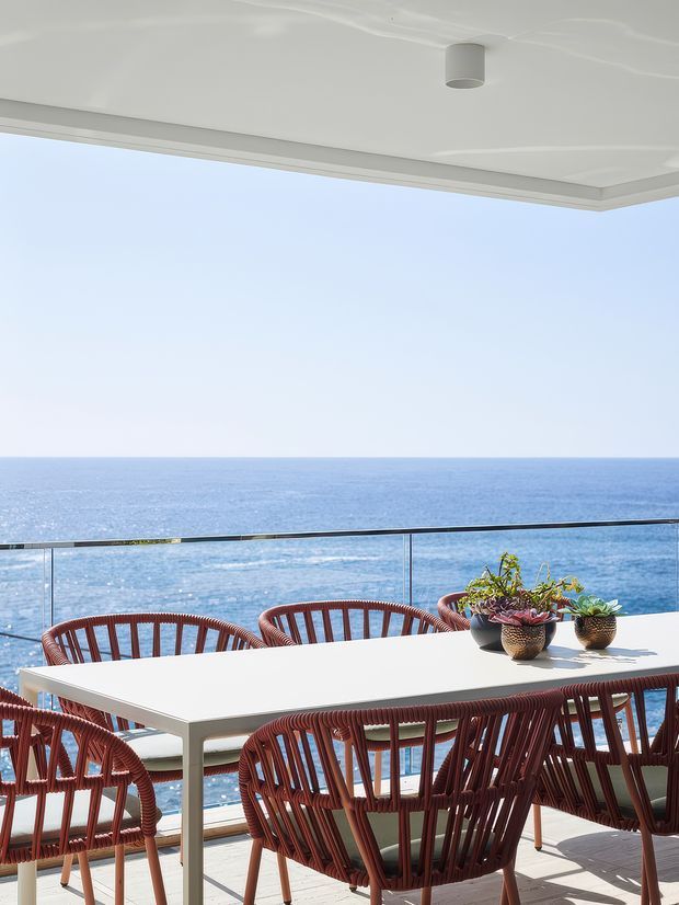 Bright coastal outdoor dining terrace overlooking the ocean, with a large white rectangular dining table, woven reddish-brown outdoor chairs, potted plants, glass balustrade, and a white covered ceiling with a simple cylindrical light. The scene has an expansive sea view and a clean, contemporary resort-like feel.