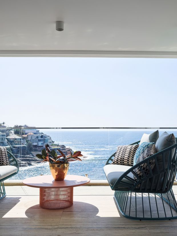 Bright coastal living area opening onto a balcony or terrace, with floor-to-ceiling glazing and a frameless glass balustrade overlooking the ocean and rocky shoreline. The space features light timber flooring, a round sculptural coffee table, dark woven lounge chairs, white cushions, and patterned textiles. Strong indoor-outdoor connection with abundant natural light and ocean views.