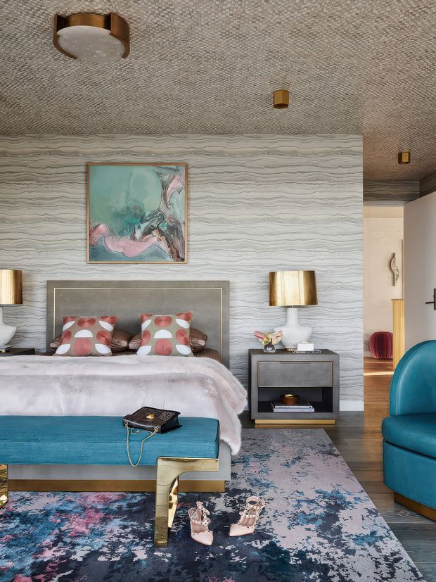 A richly layered bedroom with a bed, upholstered teal bench, bedside table, table lamps, and a bold patterned area rug. The room features a textured wave-patterned wall finish, a tightly patterned textured ceiling, colourful abstract artwork, patterned cushions, and jewel-toned furniture, creating an eclectic and luxurious atmosphere.