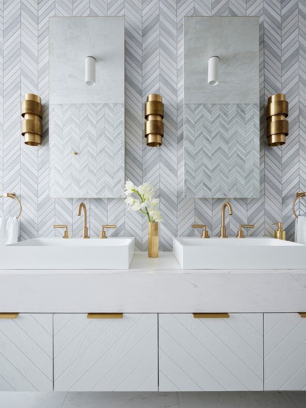 A refined double-vanity bathroom with two white rectangular vessel sinks set on a pale stone countertop. The wall is clad in a light grey-white chevron patterned finish, complemented by large rectangular mirrors, sculptural brass wall sconces, and brass tapware. A white vanity with geometric panelled cabinetry and slim brass pulls spans the lower portion of the image. The styling is symmetrical, polished, and luxurious with a soft neutral palette and warm metallic accents.