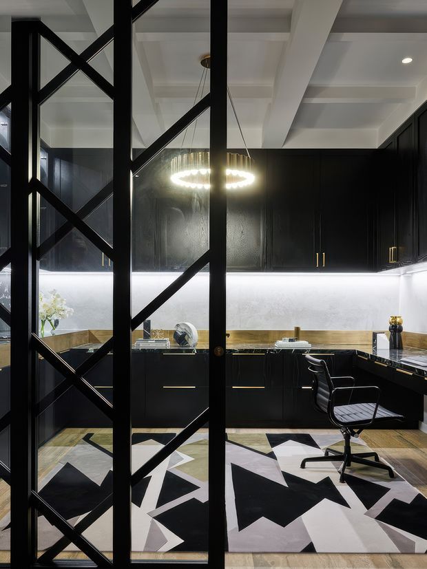 A dramatic contemporary kitchen viewed through a black metal-framed glass partition with diagonal geometric bracing. The kitchen features matte black cabinetry with brass handles, dark stone-look benchtops, a light backsplash with under-cabinet lighting, and a white coffered or beam-lined ceiling. A sculptural circular pendant hangs above the work area, while a black office chair sits on a bold geometric black, white, and grey rug over timber flooring. The space has a refined, high-contrast, art-deco-inspired atmosphere.
