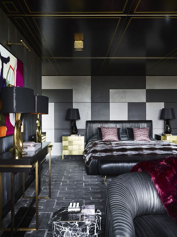 A dramatic, highly stylised bedroom with a black and charcoal palette, geometric metallic wall panelling, glossy black ceiling with gold trim, and a large ribbed upholstered bed. Matching black bedside lamps, gold-accented furniture, patterned cushions, a dark marble coffee table, and a ribbed lounge chair create a luxurious art-deco-inspired interior.