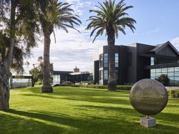 Exterior view of a large contemporary building set within a manicured lawn and landscaped garden. The dark vertically clad facade combines rectilinear volumes with a prominent gabled roof section and extensive floor-to-ceiling glazing. Tall palm trees frame the property, while a large spherical outdoor sculpture sits in the foreground.