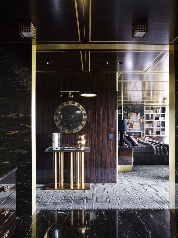 A dramatic, luxuriously styled interior foyer or hallway with dark timber wall and ceiling panelling outlined in brass trim, black marble surfaces, and a patterned grey carpet. A slim console table with brass cylindrical legs sits beneath a round decorative mirror. Through the opening, a bedroom with dark bedding, extensive built-in shelving, and artwork is visible.