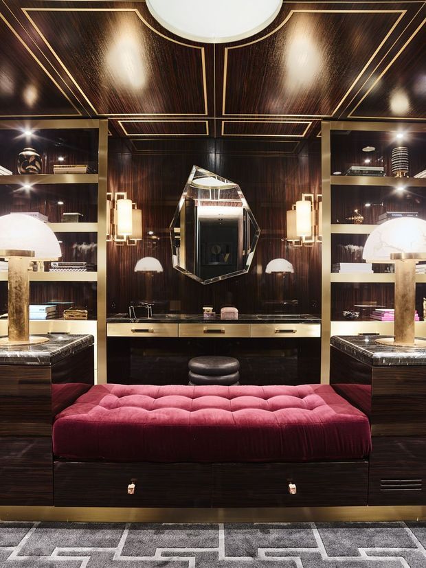 Luxurious, highly symmetrical dressing room or walk-in wardrobe with dark, high-gloss timber cabinetry and wall panelling, brass geometric inlay, mirrored shelving, integrated lighting, marble-topped side units, and a central faceted mirror. A deep burgundy tufted bench sits in the foreground over a geometric grey-and-white rug. The overall composition is richly detailed and art deco-inspired.