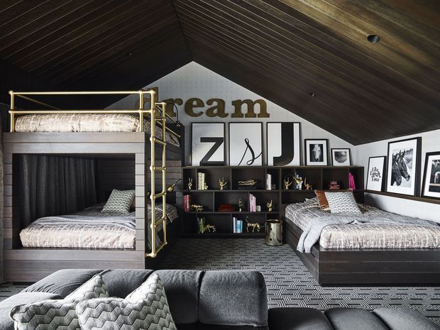 A stylish attic bedroom or kids’ bunk room beneath a dramatic sloped, dark timber-lined ceiling. The room features built-in dark timber bunk beds and a matching single bed, patterned grey bedding, open shelving with books and objects, framed artwork, and a dark sectional sofa in the foreground. Brass-toned bunk ladder and rails add a warm accent to the otherwise charcoal, black, and grey palette.