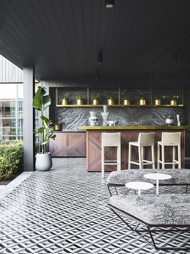 A sophisticated covered outdoor entertaining area featuring a built-in bar with a richly patterned timber front, dark stone-look backsplash, brass-toned shelving, potted plants, and white bar stools. The space has a dramatic black ceiling and geometric black-and-white tiled flooring, with a patterned outdoor lounge setting in the foreground and glazing along one side.
