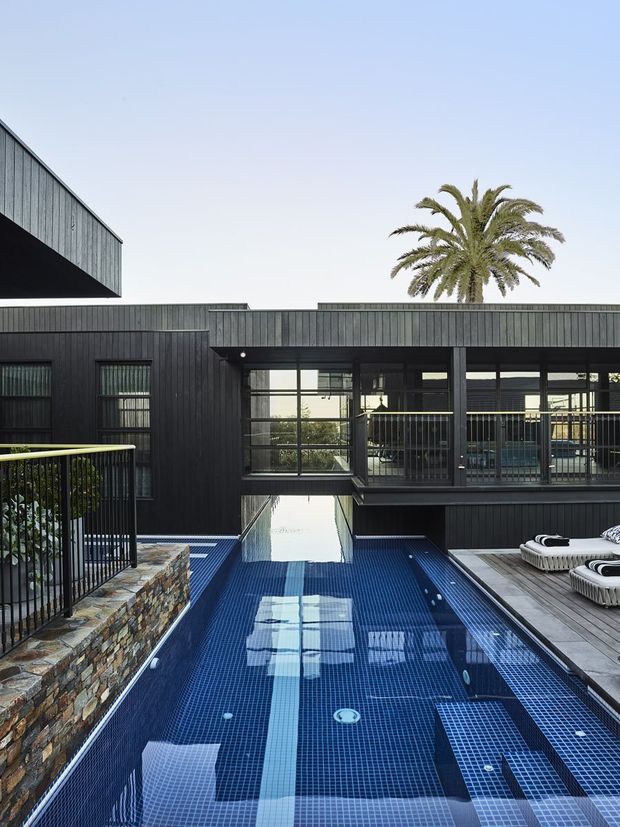 Modern dark-clad multi-level residence viewed from an elevated outdoor terrace, centered on a long blue tiled swimming pool with integrated submerged steps and a narrow reflecting-water connection beneath the building. Black metal balustrades, extensive glazing, timber decking, stacked stone retaining walls, and white pool loungers frame the scene, with a palm tree visible behind the flat-roofed structure.