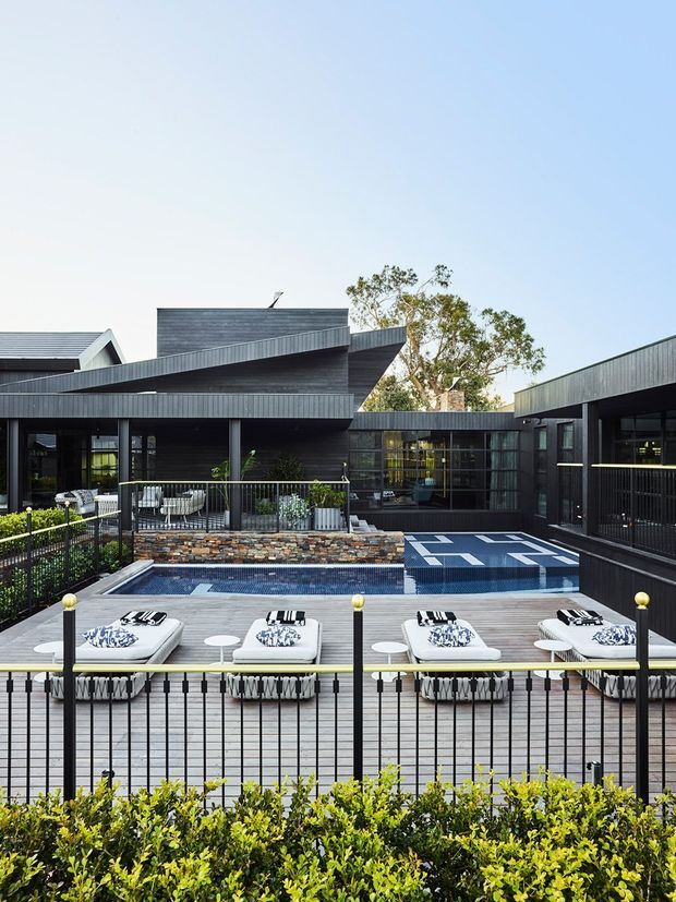 A contemporary dark-clad residential complex arranged around a raised outdoor terrace and swimming pool. The image shows angular roof forms, extensive glazing, black metal balustrades, timber decking, poolside loungers, and landscaped hedging under a clear sky.