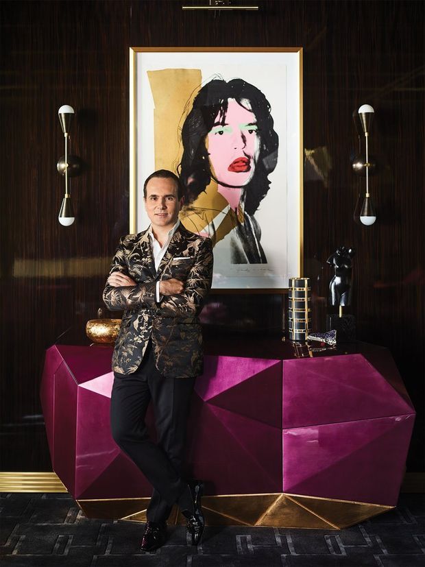 A dramatic, richly styled interior vignette featuring a man standing before a faceted magenta geometric console or bar cabinet. The room has dark glossy timber wall panelling, sculptural metallic wall sconces, a large framed portrait, metallic decorative objects, and a dark patterned carpet. The composition is highly glamorous and theatrical with jewel-toned colour and reflective finishes.