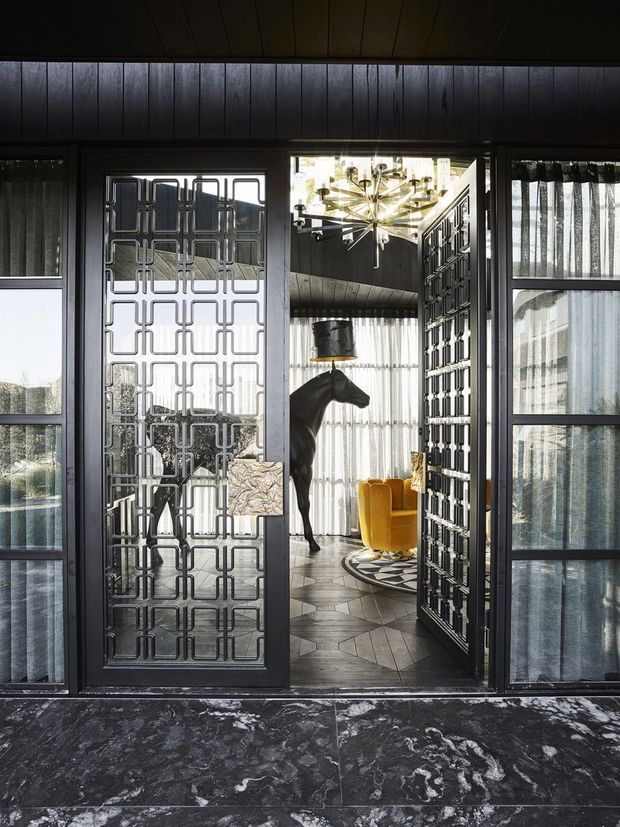 A dramatic interior view through a dark-framed glazed entry doorway into a highly styled living room. The entry features black timber wall and ceiling panelling, large dark stone floor tiles, sheer curtains, and decorative geometric metal screens in the doors and sidelights. Inside, a sculptural black horse figure, mustard-yellow lounge chairs, a patterned circular rug, and a striking starburst chandelier create a theatrical, Art Deco-inspired atmosphere.