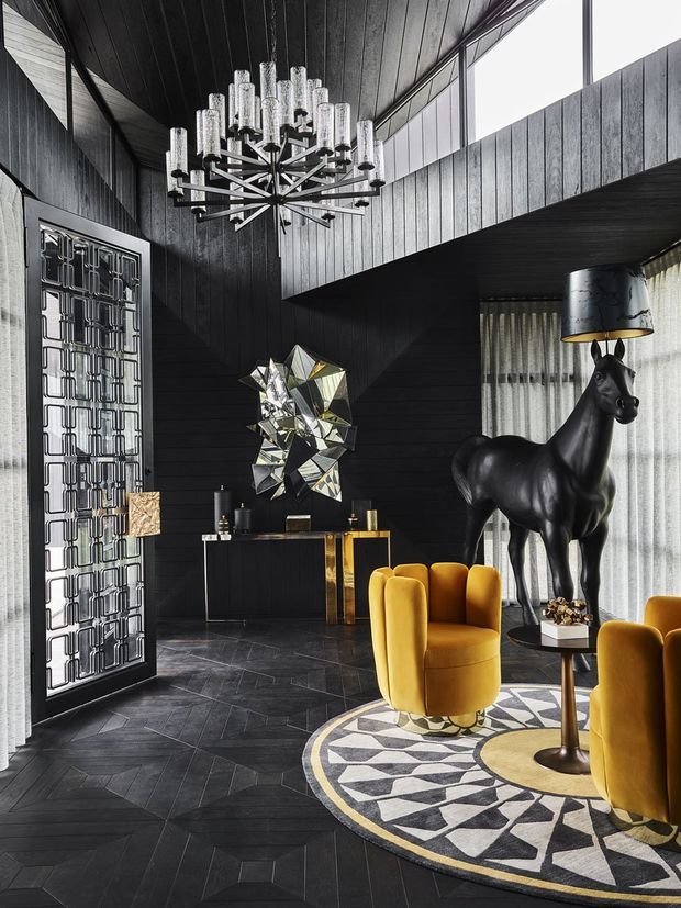 Dramatic double-height interior lounge with black timber wall and ceiling panelling, dark geometric flooring, tall clerestory glazing, and a sculptural decorative metal entry door. A striking multi-arm chandelier hangs overhead. The room features mustard-yellow upholstered lounge chairs, a black horse sculpture, a faceted wall artwork, a slim console, and a bold circular geometric rug with yellow, black, white, and grey patterning.