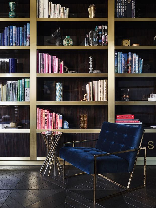 A sophisticated library or reading lounge featuring full-height dark timber shelving with brass-toned framing, densely arranged books, and curated decorative objects. A deep navy blue velvet lounge chair with a slim metallic frame sits beside a small sculptural side table. The dark geometric floor, rich materials, and dramatic lighting create a refined, luxurious atmosphere.