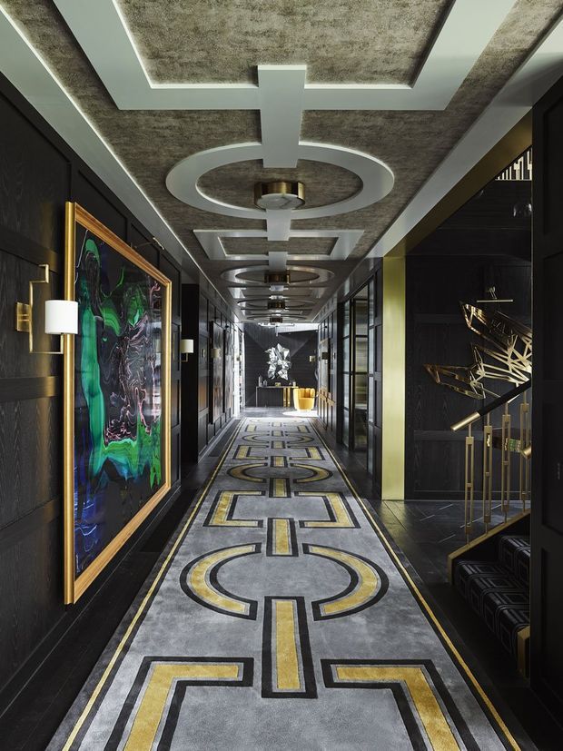 A long, highly stylised luxury hallway or gallery corridor with black wall panelling, dark timber or painted built-in cabinetry, dramatic geometric patterned carpet, and a textured concrete-look ceiling. Repeating circular and rectangular ceiling details with integrated lighting create a strong rhythm. Framed abstract artwork, sculptural objects, metallic accents, and display shelving add an art-deco-inspired, theatrical character.