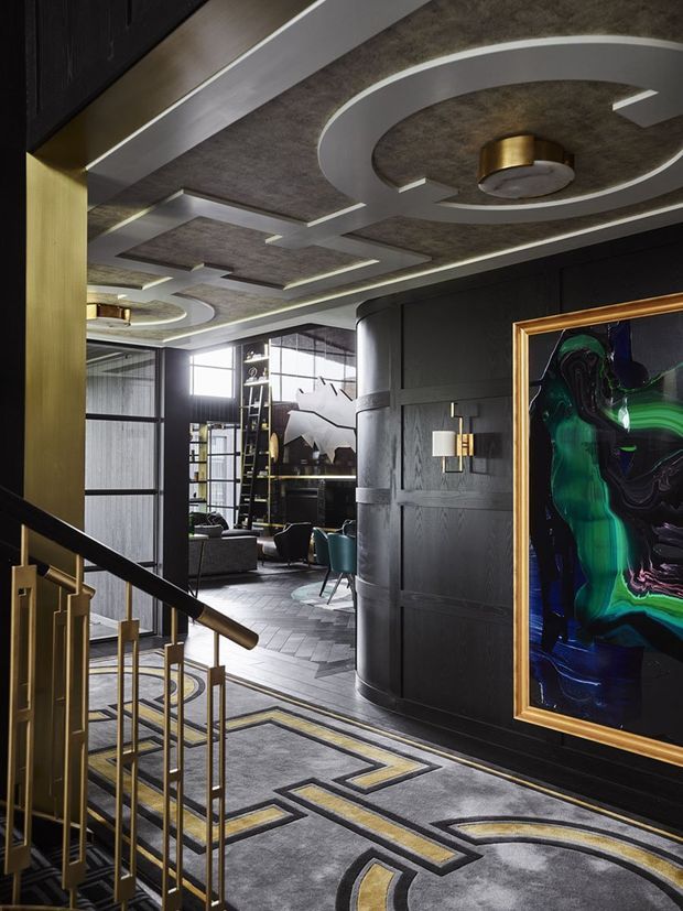 A dramatic, richly detailed interior hallway and landing viewed beside a staircase. The space features dark black wall panelling, a patterned grey and gold carpet, brass stair railings and trim, sculptural circular ceiling details with integrated lighting, and a large vivid abstract artwork in a gold frame. A lounge area with seating and shelving is visible beyond the corridor. The overall scheme is dark, luxurious and art-deco-inspired.