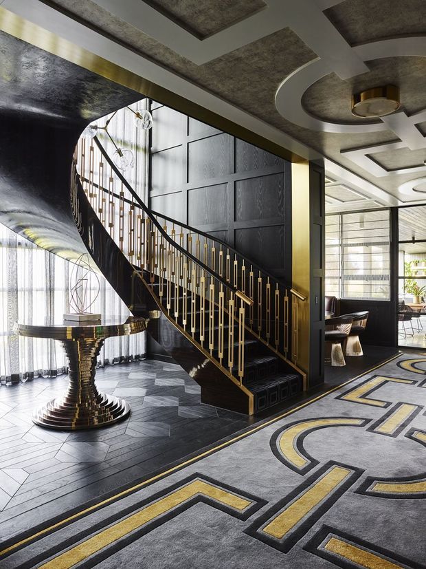A dramatic, highly detailed interior foyer featuring a sculptural sweeping staircase with black decorative wall panelling, gold-toned balustrade accents, and a reflective dark finish. The space has geometric patterned dark flooring and a large grey, black, and gold geometric rug. The ceiling incorporates layered rectangular and curved recessed panels with textured dark inserts and a central circular feature light. Full-height sheer curtains, a sculptural metallic table, and a glimpse of a dining or seating area are visible beyond the stair hall. The overall composition is luxurious, art deco-inspired, and high contrast.