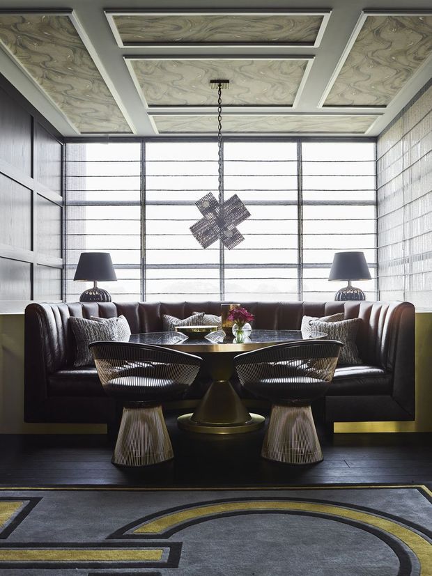 A sophisticated, symmetrical lounge or sitting area with a dark brown leather sofa, sculptural wire-frame lounge chairs, a round pedestal coffee table, matching table lamps, and a geometric patterned rug. The room features dark timber wall panelling, a large gridded window with translucent blinds, glass-block side walls, and ornate recessed ceiling panels with swirling stone-like patterns. The overall atmosphere is moody, luxurious, and strongly Art Deco-inspired.