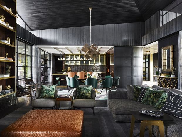 A sophisticated open-plan living and dining interior with dark timber-lined walls and ceiling, expansive black-framed glazing, a formal dining setting, and a lounge area furnished in deep charcoal and green tones. Built-in shelving, a bar or kitchen zone, sculptural pendant lighting, patterned upholstery, and a large textured leather ottoman create a dramatic, richly layered space.
