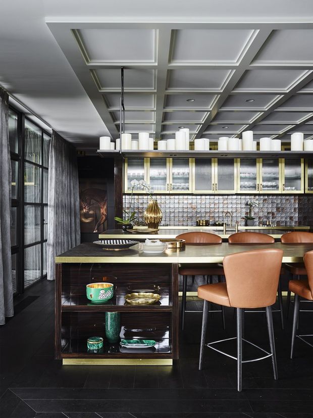 A sophisticated, dark-toned kitchen and dining space with a large island incorporating open display shelving, leather bar stools, and a metallic green-grey benchtop. The room features black cabinetry and flooring, a reflective textured tile splashback, illuminated glass-front upper cabinets with brass-toned framing, and a dramatic coffered ceiling with recessed lighting. Tall black-framed glazing and sheer curtains line one side, while decorative ceramics and a sculptural pendant-style display add visual interest.