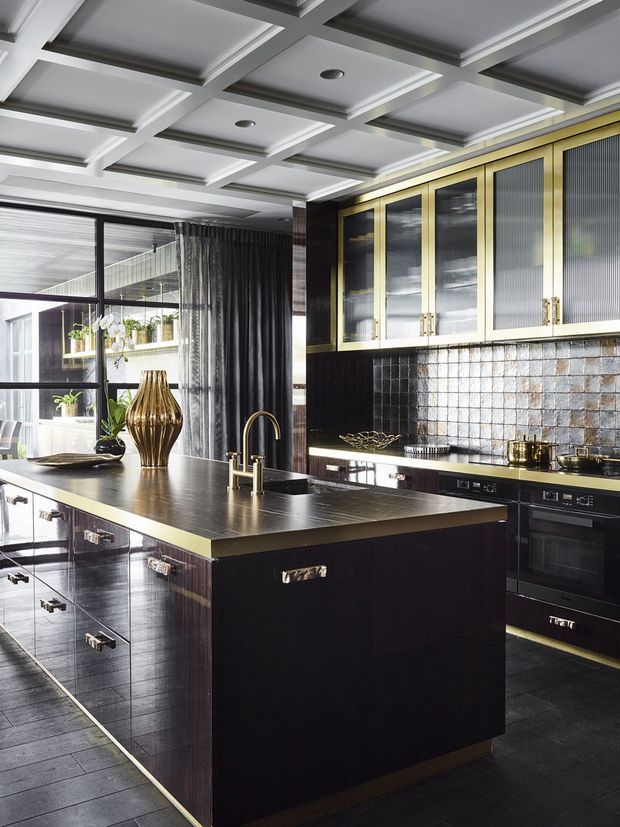 A luxurious, highly detailed kitchen featuring a large dark island with a metallic-edged benchtop, dark glossy cabinetry, brass-gold accents, glass-front upper cabinets, and a reflective metallic mosaic splashback. The room has a dramatic coffered ceiling, black-framed glazing, dark curtains, integrated appliances, and layered contemporary lighting.