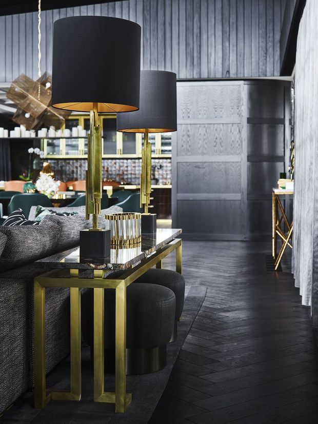 A moody, luxurious living room with a dark charcoal sofa, black cylindrical floor lamps, brass-gold metal console table, and black upholstered stools. The room features dark herringbone timber flooring, textured grey wall finishes, dark built-in cabinetry, and a sophisticated bar or kitchen area in the background.