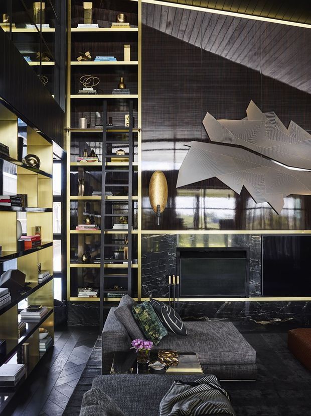 A sophisticated, dark-toned living room with glossy black marble or stone feature walls, integrated fireplace and television, full-height open shelving, and dramatic sculptural wall art. A large charcoal sectional sofa and ottoman sit on dark flooring, with brass and gold accents throughout the joinery, lighting, and decor. The room has a luxurious, high-contrast contemporary interior with reflective surfaces and carefully styled objects.