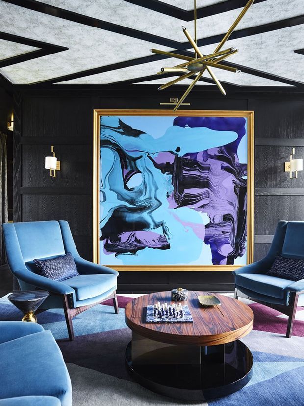 A dramatic, richly layered living room featuring deep black wall panelling, a large abstract artwork in a gold frame, sculptural brass starburst lighting, and blue velvet lounge chairs arranged around a round two-tier timber and black coffee table. A geometric blue, grey, and mauve rug adds pattern and colour underfoot.